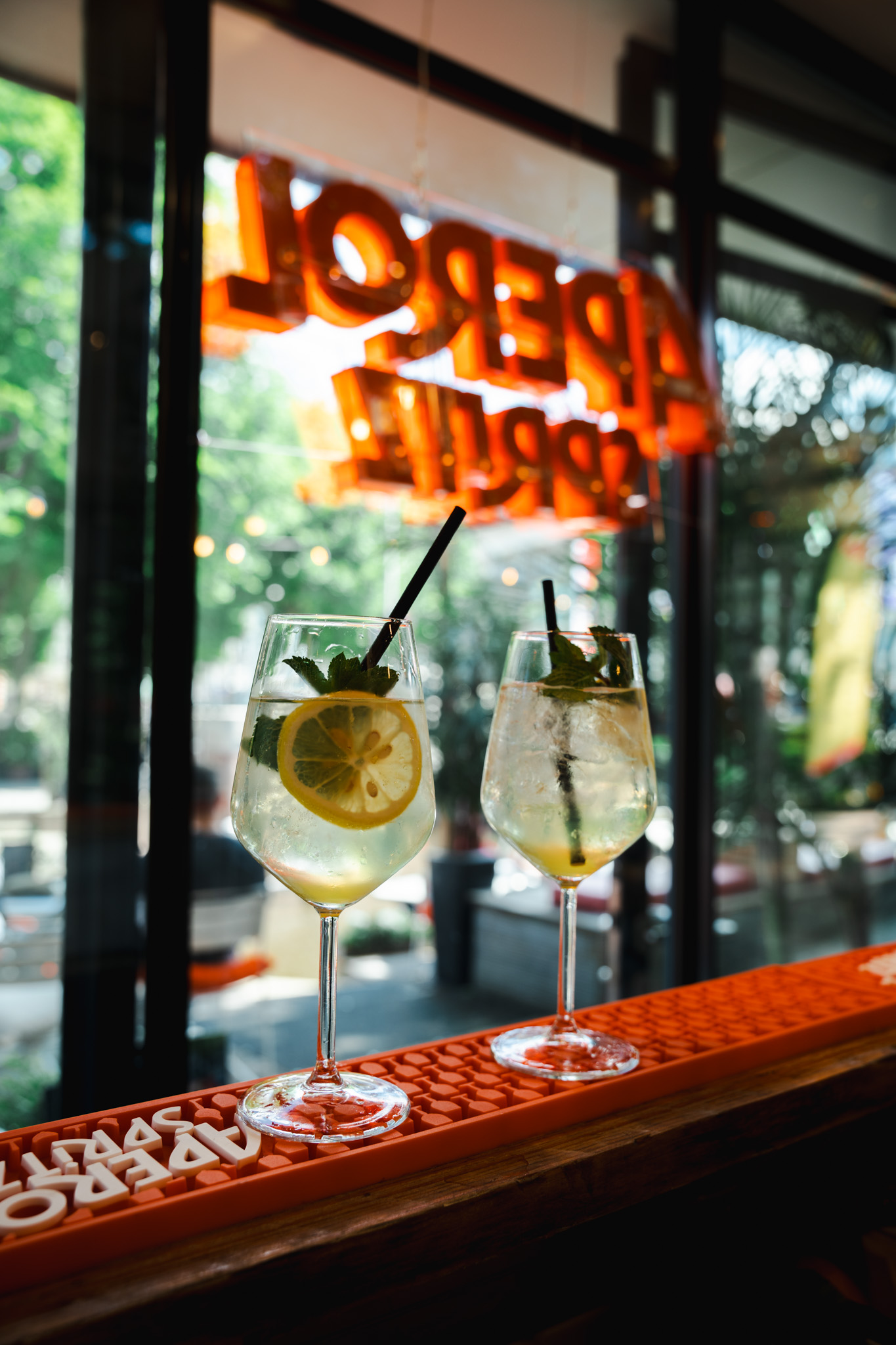Two Hugo spritzes on the bar with neon Aperol sign reflected in window